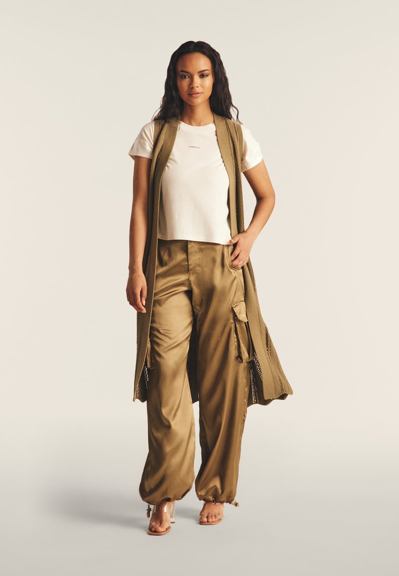 Chaleco de punto en verde oliva sobre una camiseta blanca cropped, combinado con pantalones satén en color beige con bolsillos laterales. Sandalias sencillas de tiras.