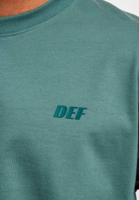 Turquoise cotton t-shirt featuring a crew neck and a textured "DEF" logo in a darker shade of turquoise on the left side.