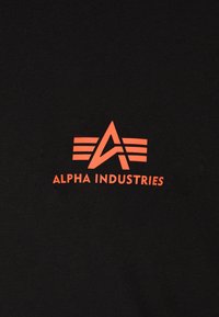 Camiseta negra con un destacado logotipo naranja de "ALPHA INDUSTRIES" con letras estilizadas y acentos a rayas. Textura de tejido de algodón suave.