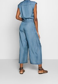 Tuta in denim con maniche corte, vita alta, gamba ampia, colore azzurro chiaro e rifinitura pulita; abbinata a sandali piatti con cinturini.