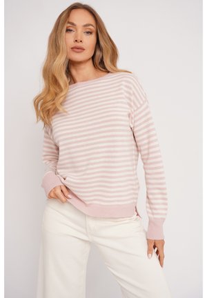 Kobieta z długimi, falującymi blond włosami, ubrana w jasnoróżowy sweter w białe paski i białe spodnie, pozująca na tle jasnego, jednolitego tła.