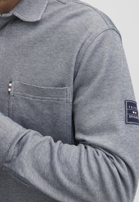 Majica z dolgimi rokavi iz teksturiranega modrega materiala. Ima žep na prsih z označevalno nalepko v črtah in povoščen kos na rokavu z informacijami o blagovni znamki.