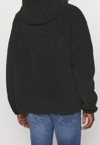 Felpa nera in pile con superficie testurizzata, ampio cappuccio e vestibilità morbida. Abbinata a jeans blu, per uno stile rilassato.