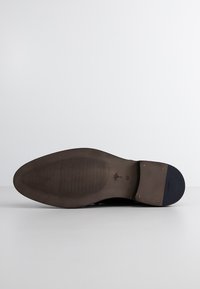 Semelle de chaussure en cuir avec un fond brun texturé, dotée d'un talon noir lisse et de détails de logo. Bout arrondi et construction robuste.