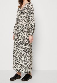 Robe maxi florale en crème avec des motifs noirs et verts, dotée d'un décolleté en V profond et de manches longues. Associée à des sabots noirs.