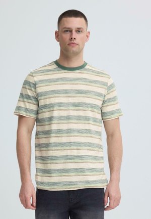 Mand vendt fremad iført en beige og grønstribet t-shirt med korte ærmer og mørke bukser mod en ensartet lys baggrund.