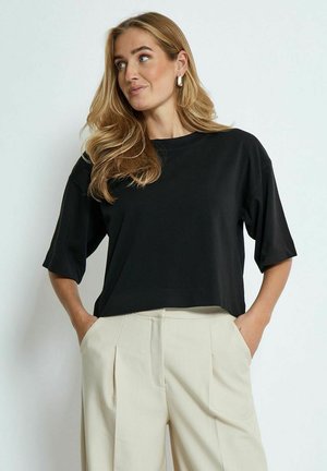 T-shirt nera cropped con vestibilità ampia e maniche corte, realizzata in tessuto morbido. Abbinata a pantaloni beige con vita alta.