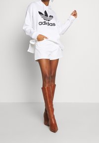 Valkoinen huppari, jossa on musta Adidas-logo, valkoiset shortsit ja pitkät ruskeat nahkaiset korkeat saappaat. Sileät pinnat ja määritellyt muodot.