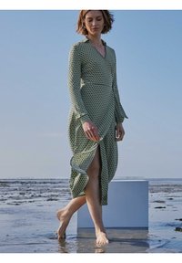 Lang groene jurk met een geometrisch patroon, met een V-hals, lange mouwen en een zijsplit, gedragen door een persoon die op nat zand loopt.