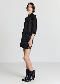 Schwarze transparente Bluse mit Kragen und Bindeband, kombiniert mit einem schwarzen Leder-Minirock und Stiefeletten mit spitz zulaufender Spitze und niedrigem Absatz.