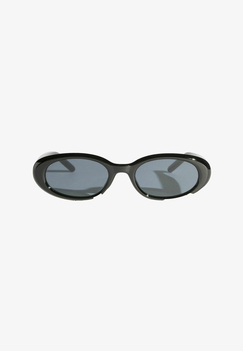 Lunettes de soleil ovales noires avec verres teintés foncés et montures épaisses brillantes sur fond blanc.