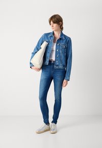 Kék farmerdzseki klasszikus dizájnnal, amelyhez slim-fit kék farmernadrág és ezüst színű sportcipő társul. Egy világos színű, texturált táskát tart.