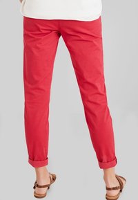 Pantalons chino en coton rouge avec une coupe ajustée, ourlets retroussés et une texture lisse, associés à des sandales plates brunes.
