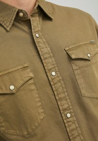 Khakifarbenes Button-Up-Hemd aus Baumwolle mit zwei Brusttaschen, Druckknöpfen und sichtbaren Nähten. Glatte Textur und lässige Passform.