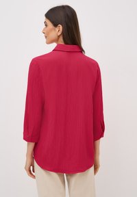 Camisa roja texturizada con cuello y mangas arremangadas, que presenta un corte relajado y un dobladillo ligeramente curvado.