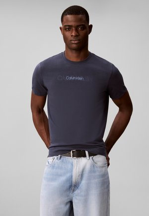 Mand iført en mørkeblå Calvin Klein t-shirt og lyseblå jeans med sort bælte står mod en ensfarvet grå baggrund.