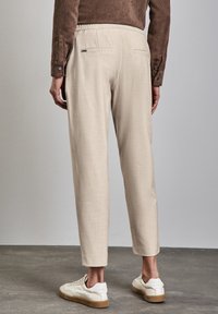 Béžové, strukturované kalhoty s elastickým pasem, zúženými nohavicemi a dvěma zadními kapsami. Nošeny s bílými teniskami na šedé podlaze.