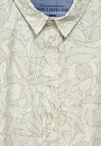 Ljusfärgad skjorta med ett tunt, lövmönster. Krage med fyra knappar framtill; mjuk tygkvalitet.