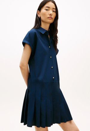 Žena u mornarskoplavoj haljini kratkih rukava s ovratnikom, zlatnim gumbima i plisiranom suknjom, stoji na jednobojnoj pozadini.