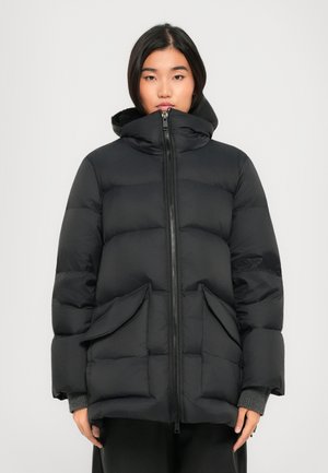 Young woman wearing a black, hooded, quilted puffer jacket with front zipper and large flap pockets, standing against a plain light background.