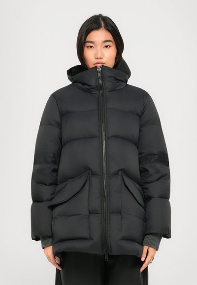 Ung kvinde iført en sort, hætte, kasseret pufferjakke med frontlynlås og store klaplommer, stående mod en simpel lys baggrund.