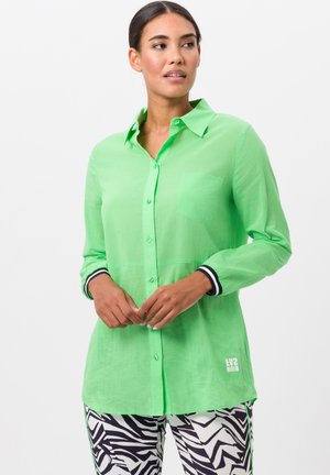 Grüne Button-Up-Hemd mit einer Tasche, aus leichtem Material gefertigt. Hat schwarz-weiße gestreifte Bündchen; kombiniert mit gemusterten Hosen.