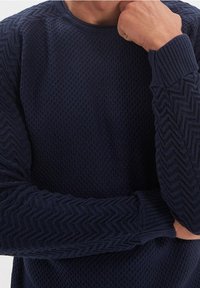 Muž na sobě má tmavě modrý pletený svetr s texturou a vzorem ve tvaru šípů na rukávech, odpočívá si bradou na pravé ruce, levá ruka zkřížená.