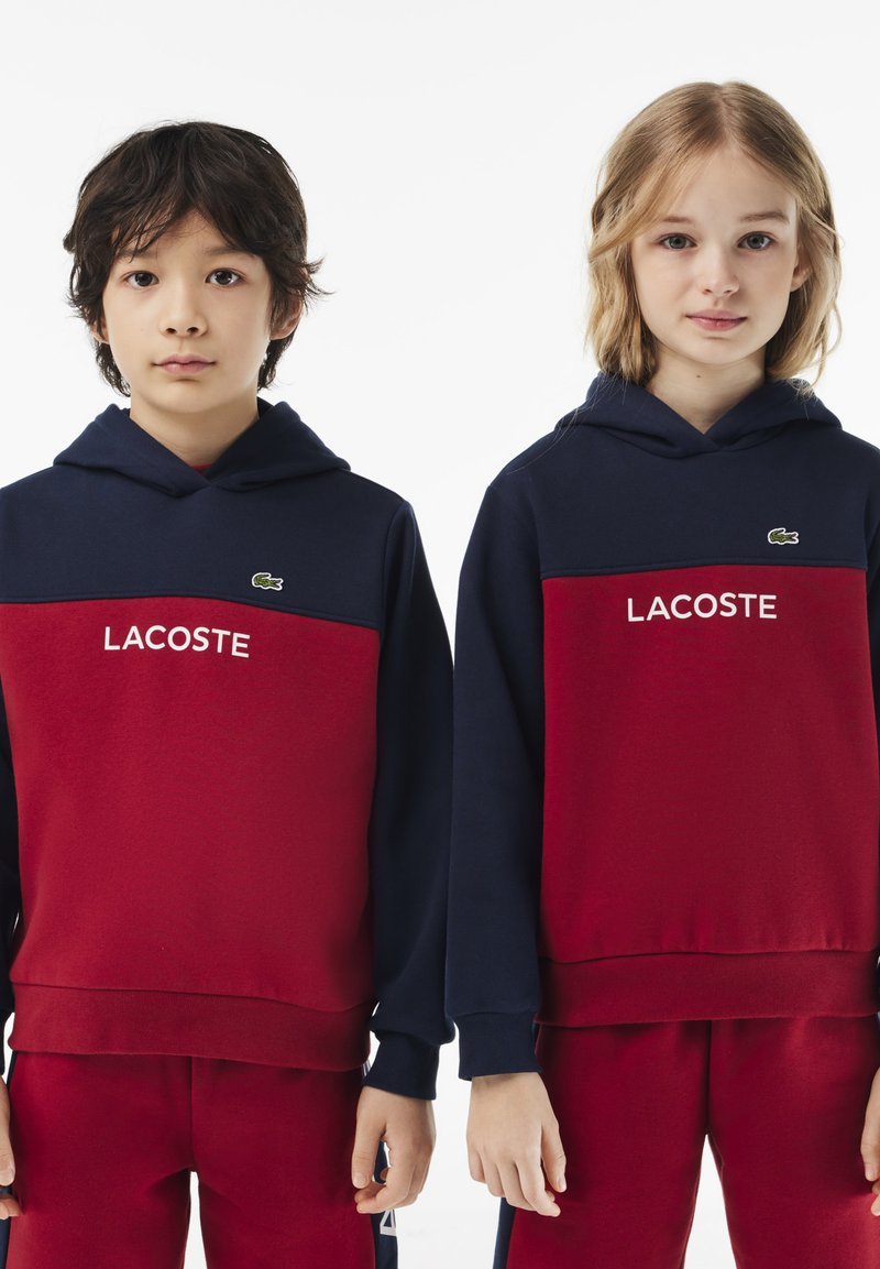 Zwei Kapuzenpullover in Navy und Rot. Beide haben das Wort "LACOSTE" in weißen Buchstaben, mit einem kleinen Logo auf der Brust.