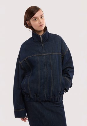 Mujer con chaqueta vaquera oversized azul oscuro de cuello alto y falda vaquera a juego, de pie contra un fondo claro y liso.