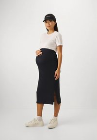 Tenue de maternité composée d'un chemisier blanc à manches courtes et d'une jupe noire moulante avec une fente latérale, associée à des baskets blanches.