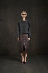 Femme portant des lunettes de soleil foncées, une veste grise à boutons avec des manches larges, une jupe midi en cuir marron avec une fente à l'avant et des chaussures sombres.