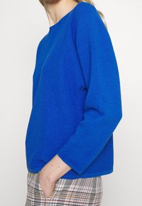 Blå ribbad tröja med rund halsringning och långa ärmar, med texturerat tyg och avslappnad passform, kombinerad med rutiga byxor.