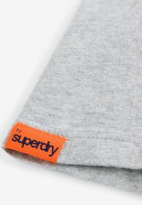 Pilkas medvilnės audinys su tekstūruota paviršiumi; turi oranžinį etiketo siuvinėjimą su užrašu "by superdry" mėlyna spalva.
