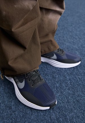 Osoba ubrana w brązowe spodnie i czarne sneakersy Nike stojąca na niebieskim, wyłożonym dywanem podłodze.