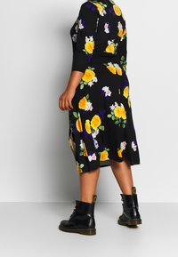 Robe noire fleurie avec des roses jaunes et blanches et des feuilles vertes, à manches trois-quart et longueur de genou, associée à des bottes noires.
