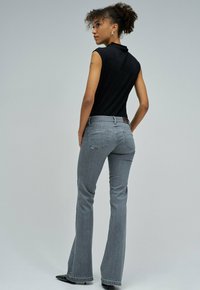 Kvinna med lockigt hår som är uppsatt, iförd en ärmlös svart topp, grå vida jeans och svarta spetsiga skor, står på sidan.