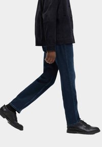 Mörkblå jeans med rak passform, kombinerade med en svart mockajacka och svarta snörskor, visade i en gående position.