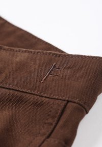 Brunt stof med en tekstureret overflade, prydet med et broderet emblem "F" i en kontrasterende tråd. Inkluderer synlige stinger langs kanten.