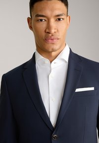 Blazer blu navy in tessuto testurizzato, chiusura a due bottoni, rever a punta e fazzoletto bianco nella tasca. Sotto indossa una camicia bianca impeccabile.