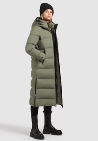 Manteau long en duvet vert olive, avec capuche, fermeture éclair à l'avant, poches latérales et texture matelassée. Porté avec des bottes noires et des leggings.