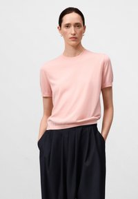Maglietta lavorata a maglia corta di colore rosa chiaro con scollo tondo, abbinata a pantaloni plettati scuri. Texture liscia, design semplice, senza motivi.
