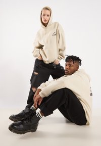 To modeller har på seg store beige hettegensere og svarte slitte jeans. Den mannlige modellen har svarte snørebukser med gule detaljer.