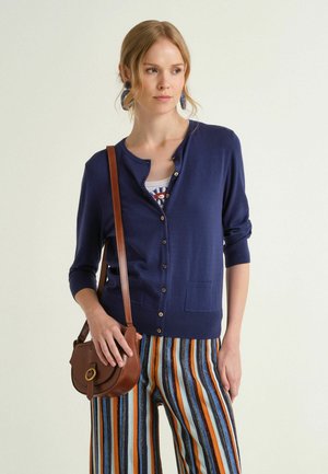 Donna che indossa un cardigan blu navy con bottoni, pantaloni a righe multicolore, orecchini blu e una borsa a tracolla marrone, che guarda di lato.