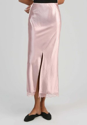 Femme portant une jupe midi en satin rose pâle avec une fente à l'avant et un ourlet en dentelle, associée à des ballerines noires à enfiler sur un fond uni.