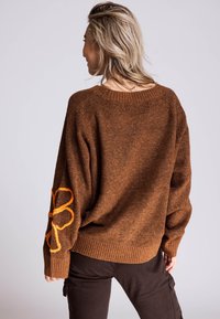 Brauner, strukturierter Strickpullover mit lockerem Schnitt, versehen mit einem kontrastierenden orangefarbenen, bestickten Blumenmotiv am linken Ärmel.