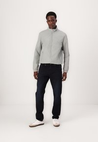 Lys grå bomberjakke med høy krage og sidelommer, paret med mørkeblå jeans. Skoene er hvite med beige detaljer.