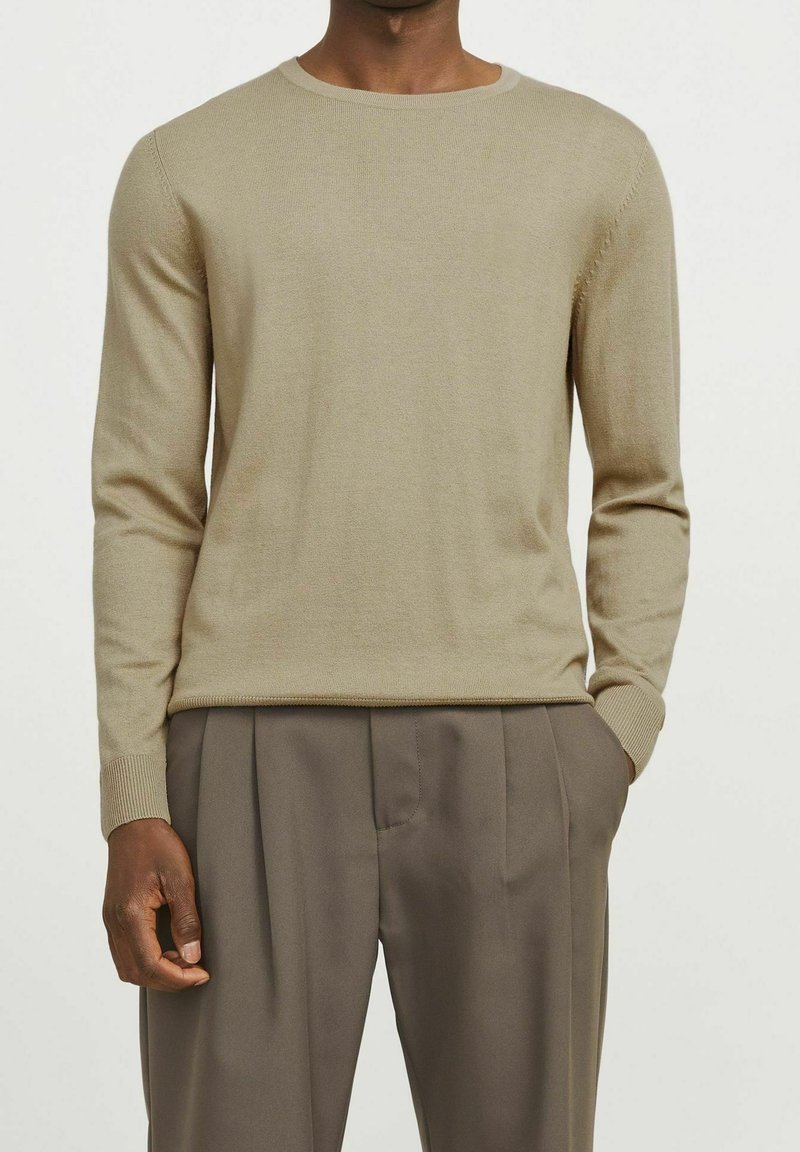 Homme portant un pull beige à manches longues et col rond, et un pantalon marron ample à plis, debout avec une main dans la poche.
