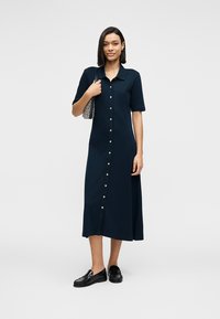 Μπλε ναυτικό μεσαίου μήκους φόρεμα με κοντά μανίκια, κουμπιά μπροστά και γιακά. Υφασμάτινη υφή με χρυσούς κουμπιά και ευθεία γραμμή. Φοράει μαύρα μοκασίνια.