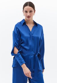 Blusa in satin blu royal con scollatura a V, maniche lunghe con spacchetti e cintura legata in vita. Tessuto morbido e design elegante.