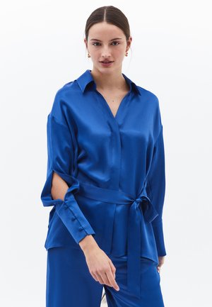 Blouse en satin bleu royal avec un décolleté en V, des manches longues avec des fentes et une ceinture nouée à la taille. Texture lisse et design élégant.